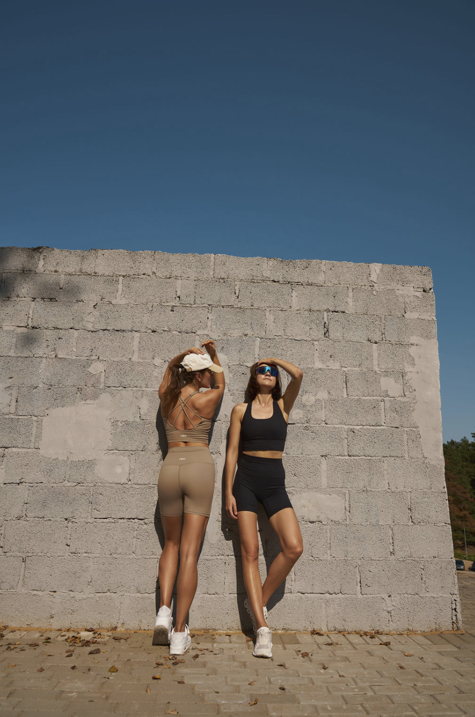 Two women posing in active wear against a stone wall with a clear blue sky.