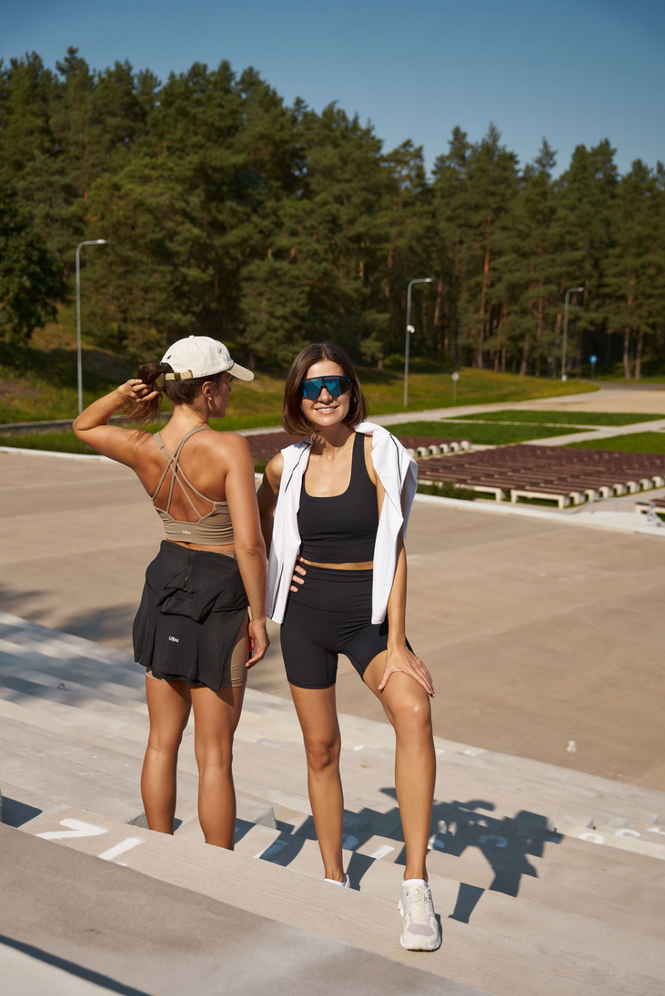 Two women in athletic Tobu Pace wear standing outdoors with trees in the background. Stretching before the exeercise 