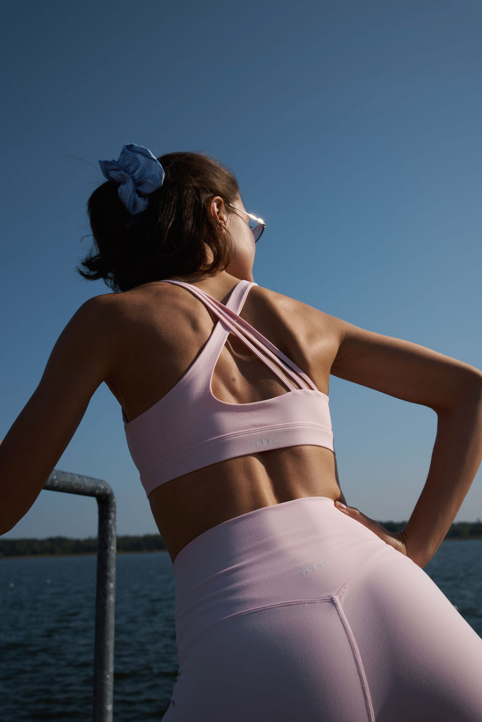 Woman in athletic wear: light pink top and light pink leggings, standing by a body of water with a clear blue sky.